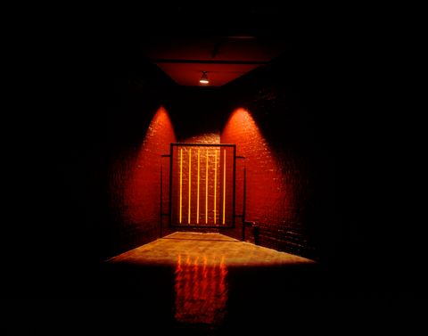 Installation. In an otherwise dark room, a spotlight creates a pool of light around a gate-like sculpture tucked into the corner of brick room, which appears to be painted red. The vertical bars of the sculpture are yellow and the framework black.