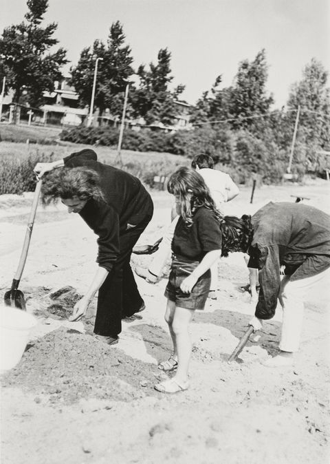 Black and white photograph. Two adults and two children dig with shovels in a field of dirt. Trees line a grassy lawn in the background.