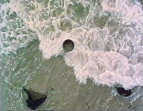 Film still. We look down onto breaking surf. A precise dark circle and two dark rocks break up the rippling sage-green water and white foam.