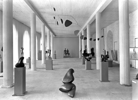 Installation photograph. This black and white photograph shows about twenty sculptures arranged mostly on plinths, though at least one is on the floor, in a hall-like, columned room.