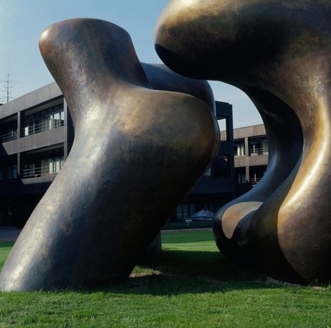 Sculpture. Two rounded, abstract forms sit on a grassy lawn in front of and nearly as tall as a three-storey building. One rounded form curves up and over the other. The surface of the bronze is darkened in some areas and matte gold in others.
