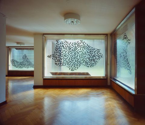 Installation photograph. Three windows in an interior space are overlaid with textured but translucent plastic. Slightly curving black lines, like bits of a circle, are gathered in abstract fields.