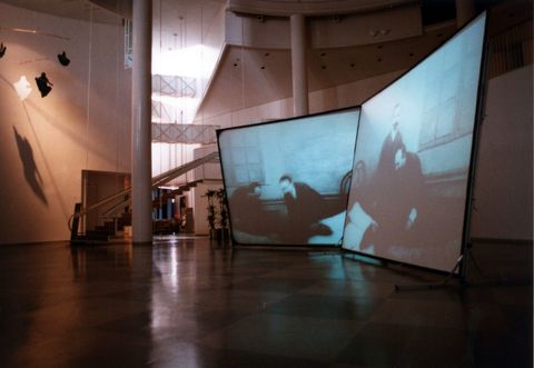 Installation photograph. Two screens tip towards or away from the viewer in an atrium-like space. Both screens show black and white stills of two pale-skinned men in dark suits.