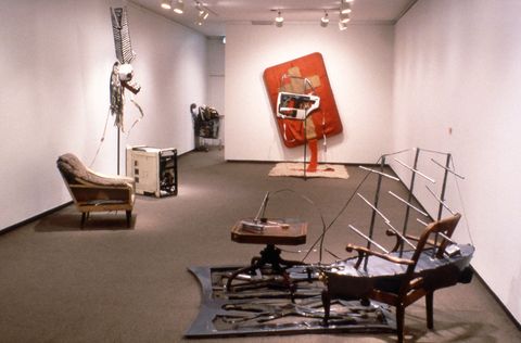 Installation photograph. Six sculptures incorporate some familiar objects, like an armchair next to an old-fashioned washing machine and a shopping cart in the next room. Some are surreal, like a chair whose seat has been replaced with a model of a boat. Another is abstract.