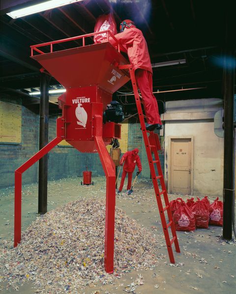 Installation photograph. A mannequin dressed in red stands near the top of a ladder and empties a red bag into a shredder, which is about two storeys high. A mound of shredded material sits under the machine. More red bags are tied to the side, and another red-clad mannequin sweeps in the background.