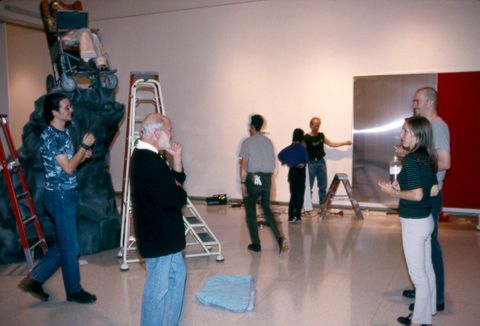 Colour photograph. Seven people stand or walk through a gallery. Ladders and step stools are set up near sculptures, which are being installed.