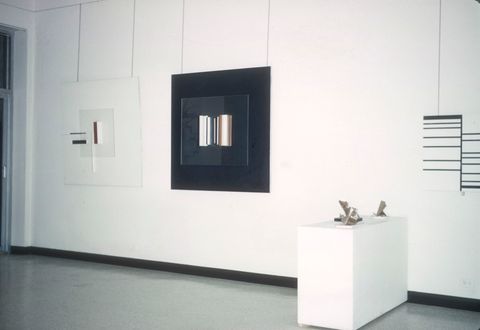 A gallery display featuring three framed minimalist artworks on the wall and a pedestal with small sculptural pieces. The left artwork is a translucent, layered composition with abstract geometric elements in black, white, and red. The center artwork is framed with a black square and features vertical bars in metallic and neutral tones, creating a reflective effect. The right artwork is composed of black horizontal lines arranged in a grid-like structure on a white background. In front of the artworks, a white pedestal holds small, abstract sculptures with metallic finishes, showcasing sharp and angular designs. The clean and bright gallery space enhances the modern aesthetic.
