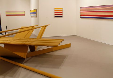 Installation photograph. An abstract sculpture made up of two long tubes balanced across V-shaped metal panels, all painted harvest yellow, is displayed in a gallery hung with horizontal, striped paintings.