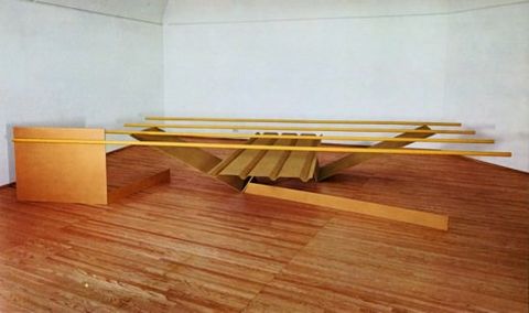 Sculpture. An abstract sculpture made of harvest-yellow, metal tubes and panels sits at the end of a gallery room with white walls and a wood floor.