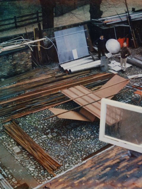 Colour photograph. We look down into a courtyard where a tawny-brown, abstract sculpture is set up. Two long tubes appear balanced across flat and V-shaped panels. Two people at the far side of the courtyard.