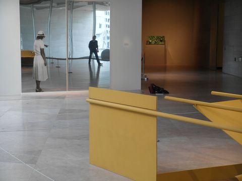 Installation photograph. The corner of an abstract sculpture edges into this photograph. A flat metal panel and three pipes, all harvest yellow, angle towards a work of art made of two rectangular pieces of reflective metal hanging on the wall opposite us. A woman wearing a white dress and pointing is screen printed onto the left of two reflective panels. A person occupying the museum space is visible in the reflection.