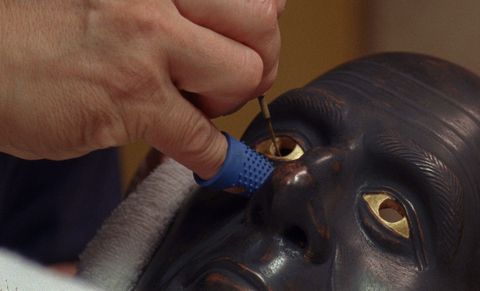 Film still. Close up of a hand working on a mask. The mask is bronzed black, and a tool is held in the cavity left for the iris. The hand has a blue rubber thimble on the pinkie finger.