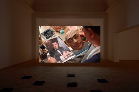 Installation photograph. A film is projected at the far end of a dim gallery. In it, a person holds a mask in one hand and a photograph, both of Sean Connery, in the other.