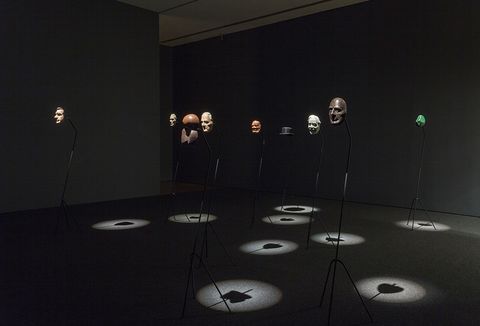 Film still. Five masks, a head-sized abstract sculpture, a top hat, and a green form are on stands in a darkened gallery. Each is spotlit from above.